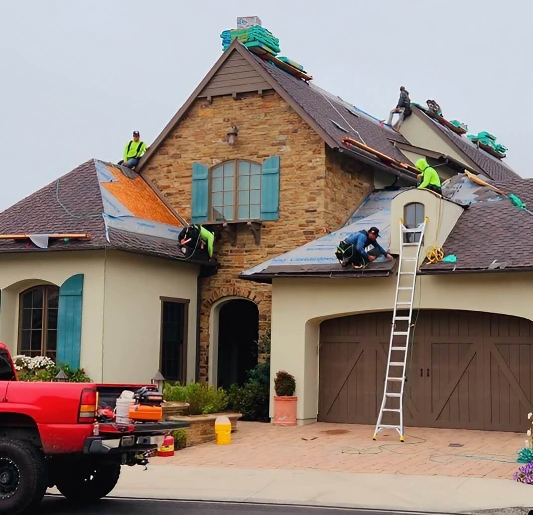 A crew of roofers in bright green vests works on a residential roof, with equipment and materials scattered around a red pickup truck.