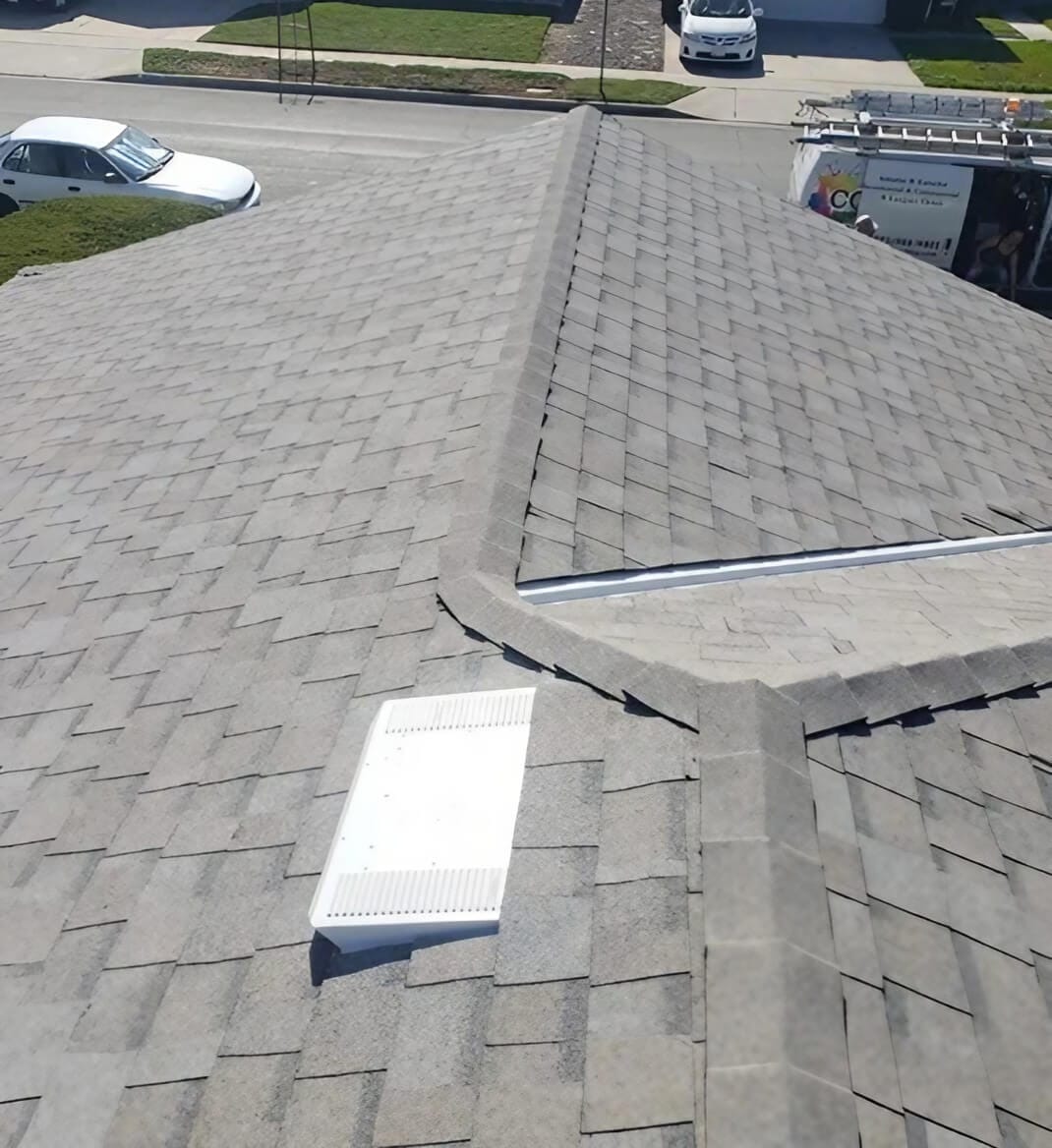 Aerial view of a gray shingled roof with a white vent on a sunny day, showcasing a well-maintained, pitched design.
