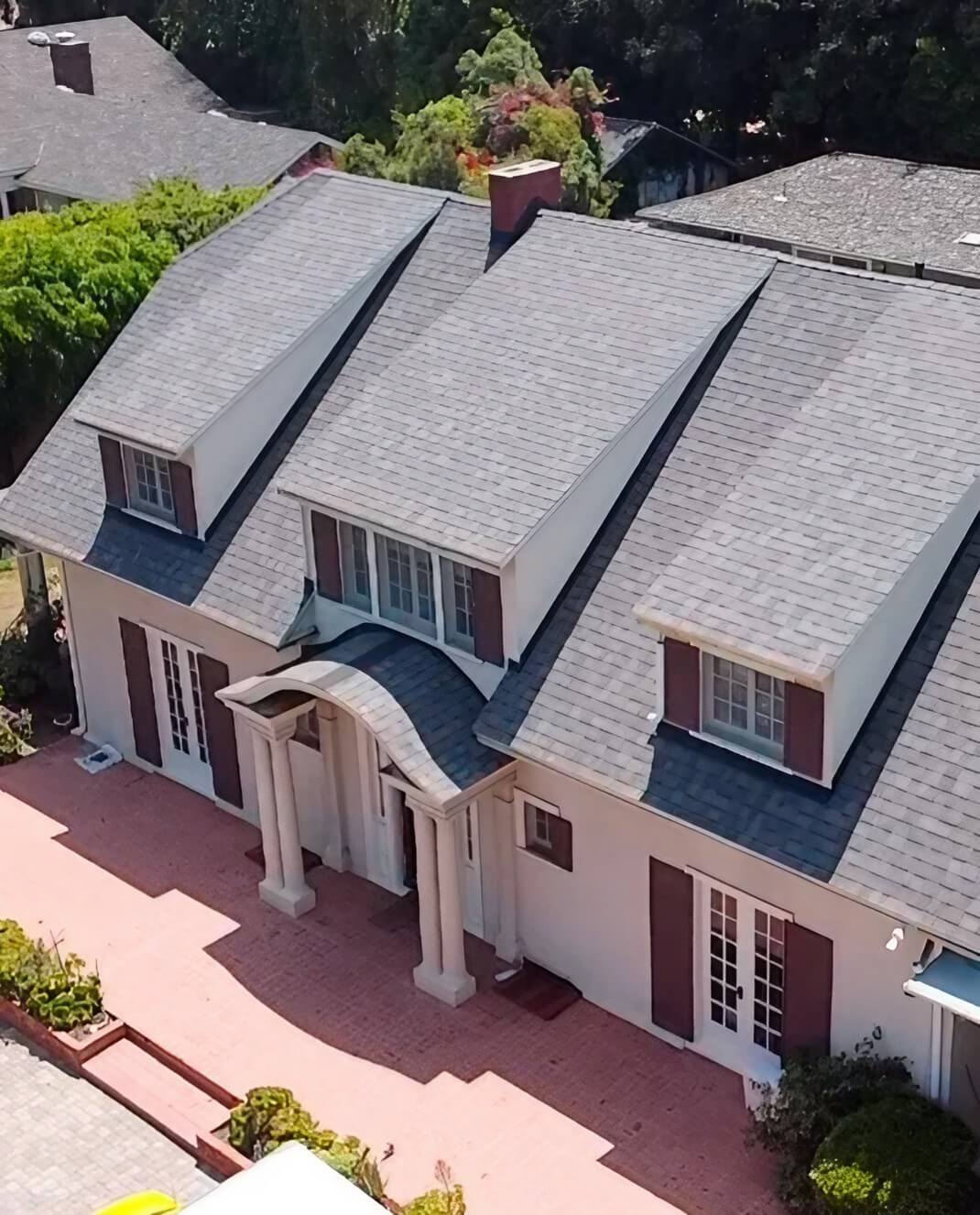 Aerial view of a classic two-story house with a grey roof, white columns, and a red brick walkway, surrounded by lush greenery.