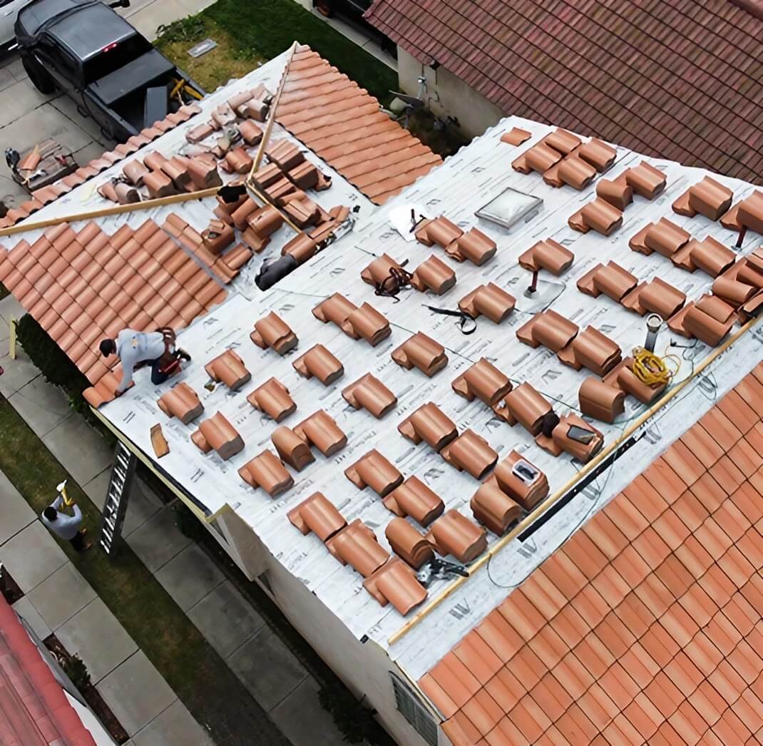Aerial view of a roof being tiled, with workers laying terracotta tiles and tools scattered across the surface.