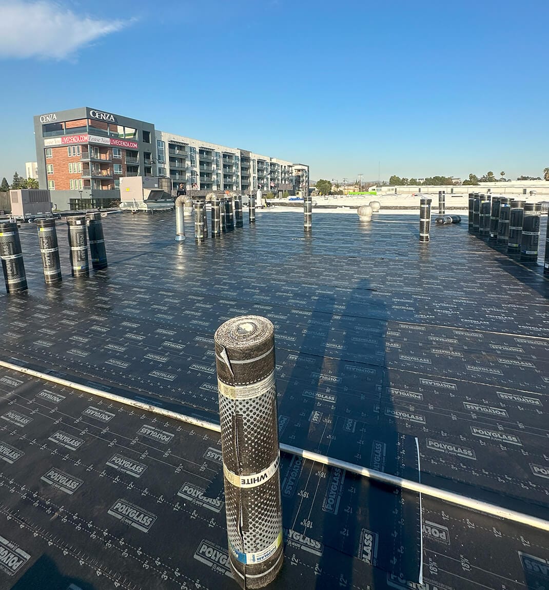 Flat rooftop covered in black waterproofing material, with rolls neatly arranged and a modern apartment building in the background.