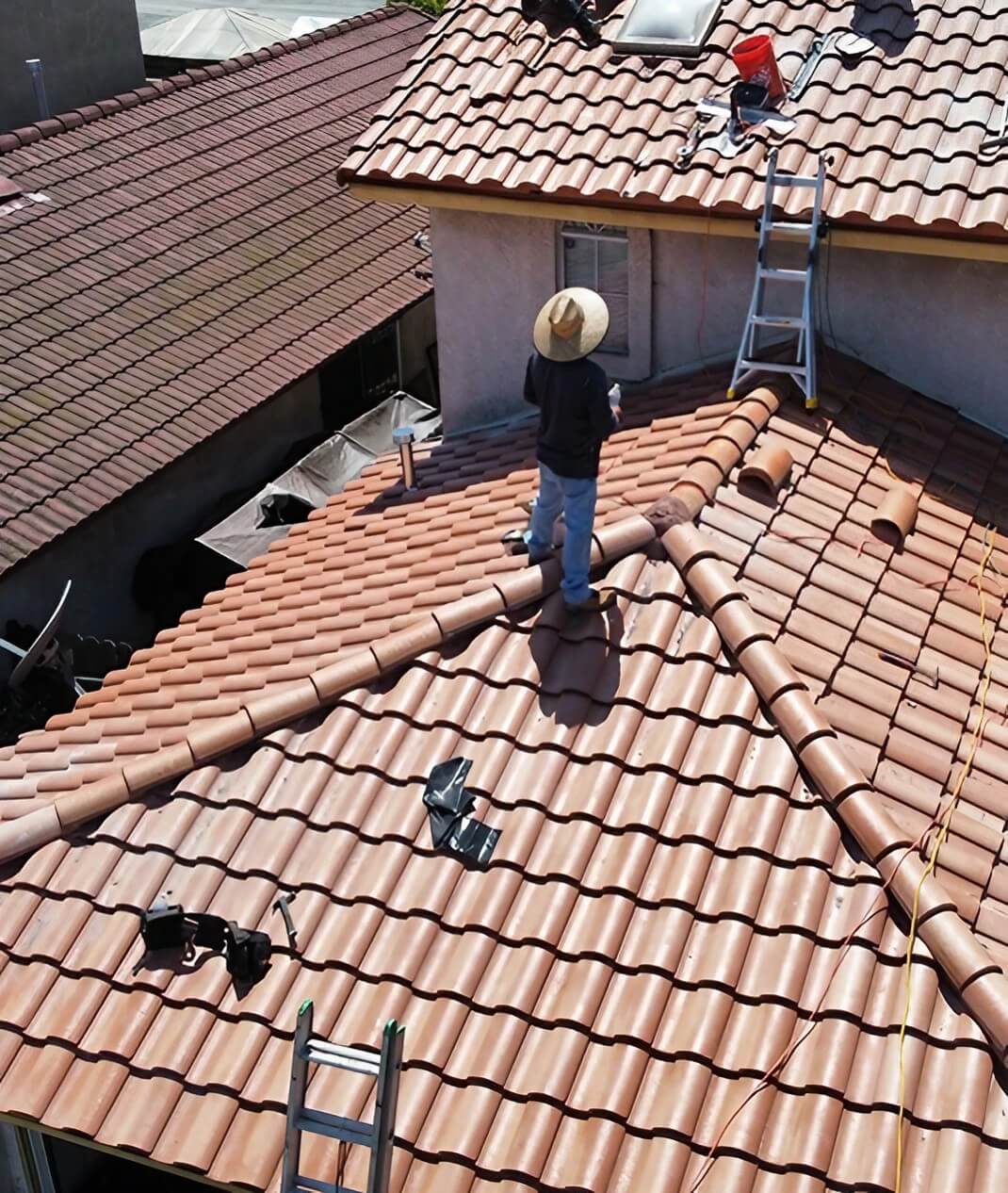 Two workers repair a tiled roof under the sun, with tools and a ladder nearby, showcasing a busy construction scene.