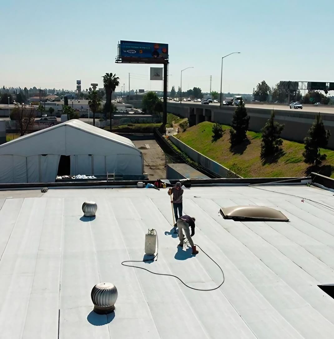 Two workers repairing a flat rooftop under a clear sky, with a large billboard and a tent in the background.