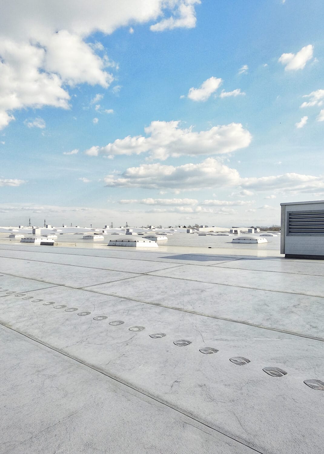 View from a rooftop with a bright blue sky, scattered clouds, and a flat, white rooftop landscape with ventilation units.