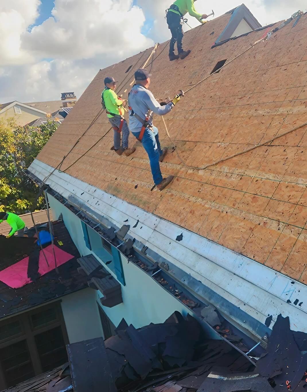 Workers in safety gear are installing a new roof, with debris scattered on the sloped surface and tools scattered around the site.