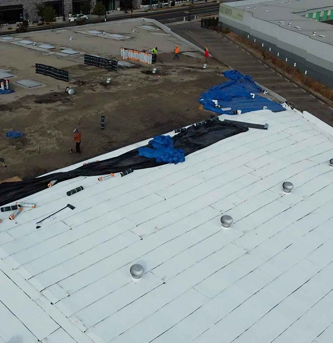 Workers on a partially completed white roof, with blue tarps and construction materials scattered around a commercial building site.